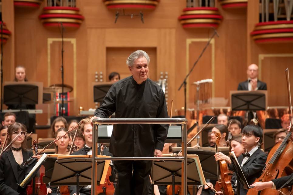 A male conductor, wearing smart black attire, standing in the conductor's podium smiling at the audience, with musicians sitting behind him.